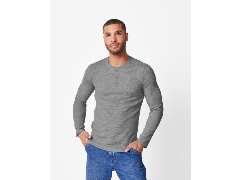 Mann in grauem Langarmshirt und blauen Jeans, mit Händen in den Taschen.