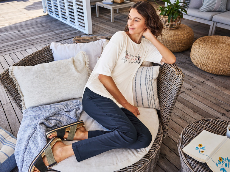 Eine Frau trägt ein weißes T-Shirt mit Anker-Druck und schwarze Hosen, während sie auf einem Rattanstuhl sitzt.