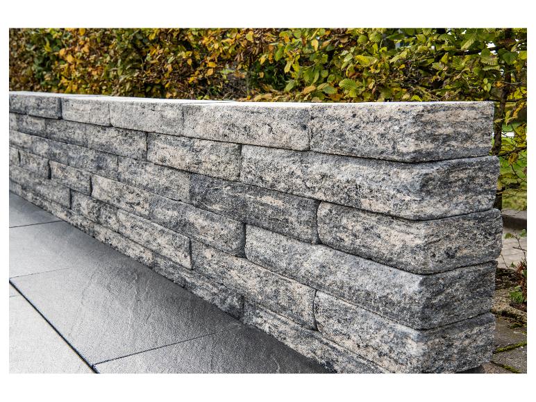 Stützmauer aus grauen und schwarzen Steinblöcken mit grünem und gelbem Laub im Hintergrund.