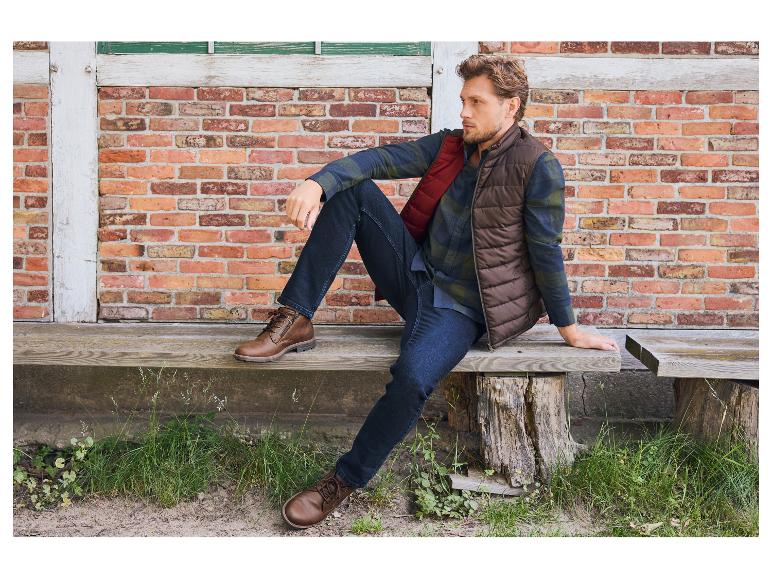 Mann in brauner Weste, kariertem Hemd und Jeans sitzt auf einer Holzbank.