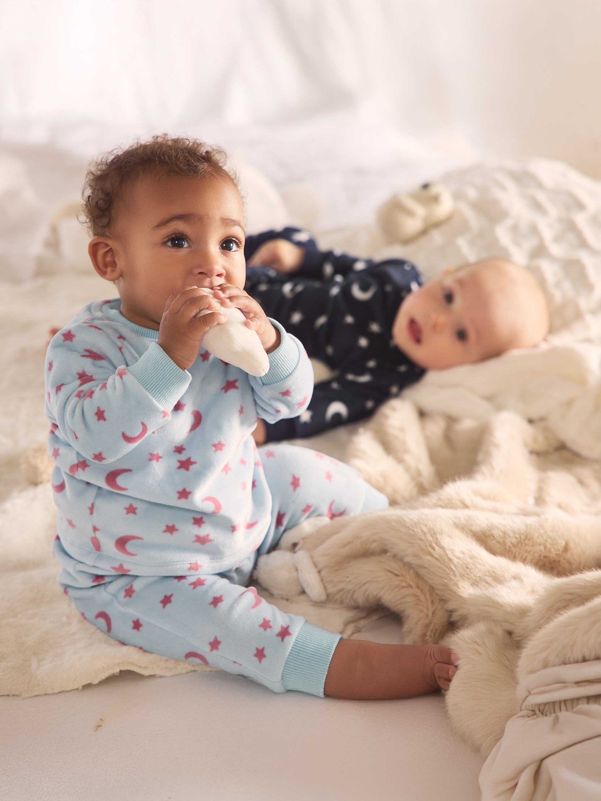 Zwei Babys in Stern- und Mondpyjamas, eines sitzend, das andere liegend auf einer weichen Decke.