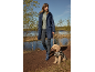 Frau in marineblauer Regenjacke und Gummistiefeln geht mit Hund in Leopardenjacke am See spazieren.