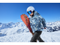 Lächelnde Frau in Skijacke und Skihose mit einem Snowboard in den Bergen.