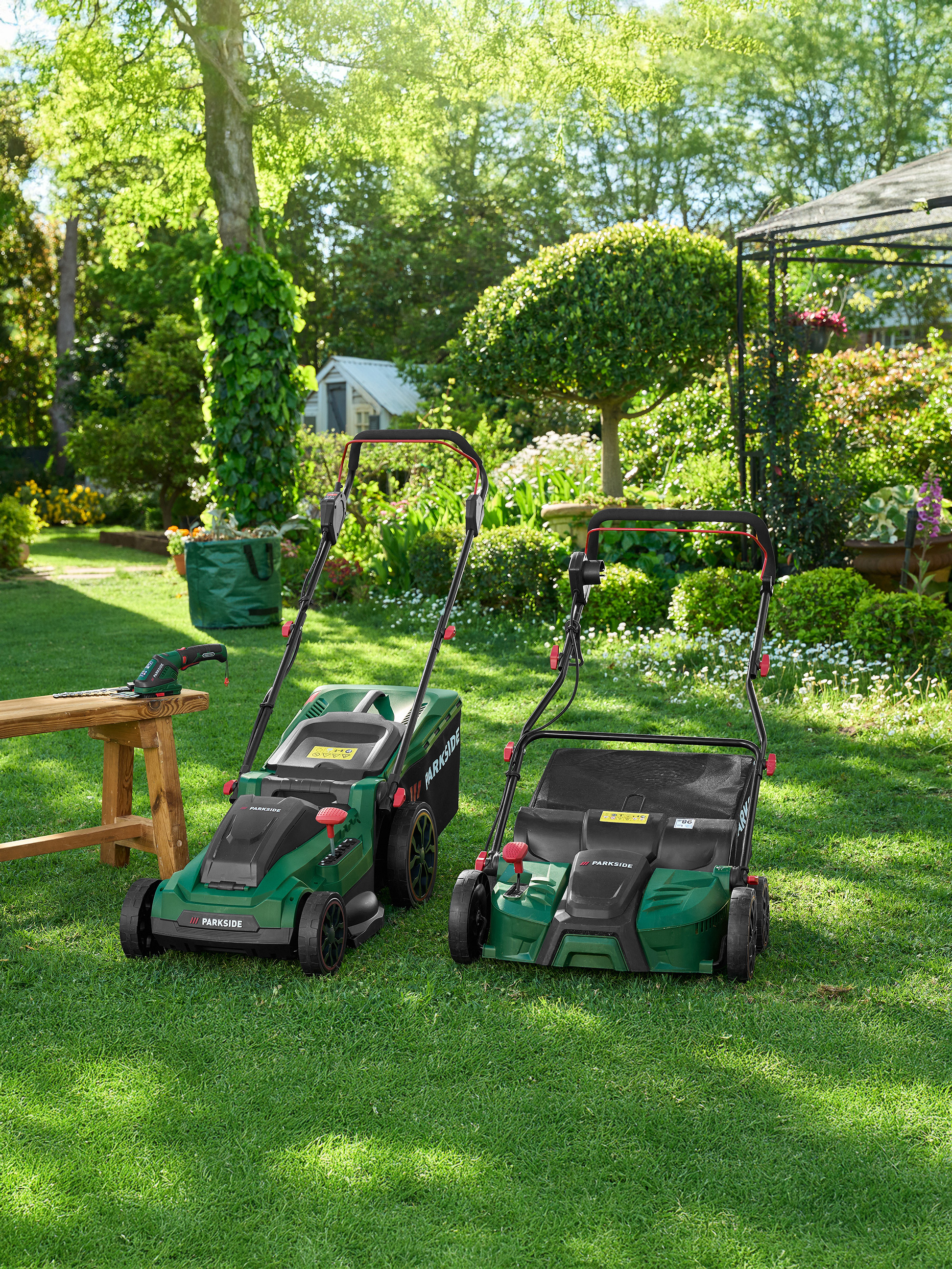 Zwei grüne Parkside Rasenmäher und Gartengeräte auf einem Rasen im Garten.