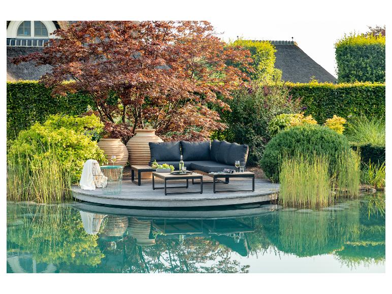Gartenmöbel auf einer Holzterrasse am Wasser, umgeben von üppigem Grün.