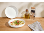 Nudelsalat in einer weißen Schüssel, mit einer Olivenölflasche und einer Reibe.