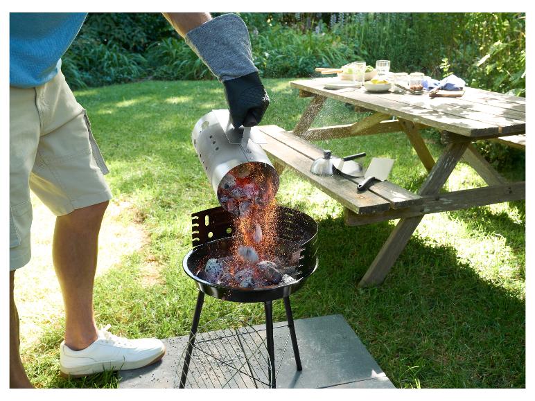 Mann schüttet glühende Kohlen in einen Holzkohlegrill im Garten, Picknicktisch im Hintergrund.