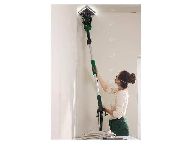 Frau mit Schutzbrille und Maske schleift Wand mit Teleskopschleifer.