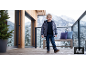 Junge in Daunenjacke und Jeans steht auf Holzterrasse mit verschneiten Bergen im Hintergrund.
