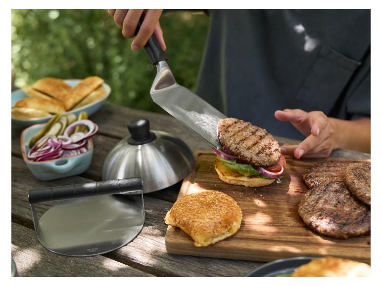 Person legt Burger mit Spatel auf Brötchen, mit Grillzutaten auf dem Tisch.