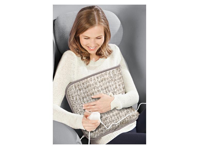 Eine Frau auf einem Stuhl lächelt, während sie ein beiges elektrisches Heizkissen auf ihrem Schoß hält.