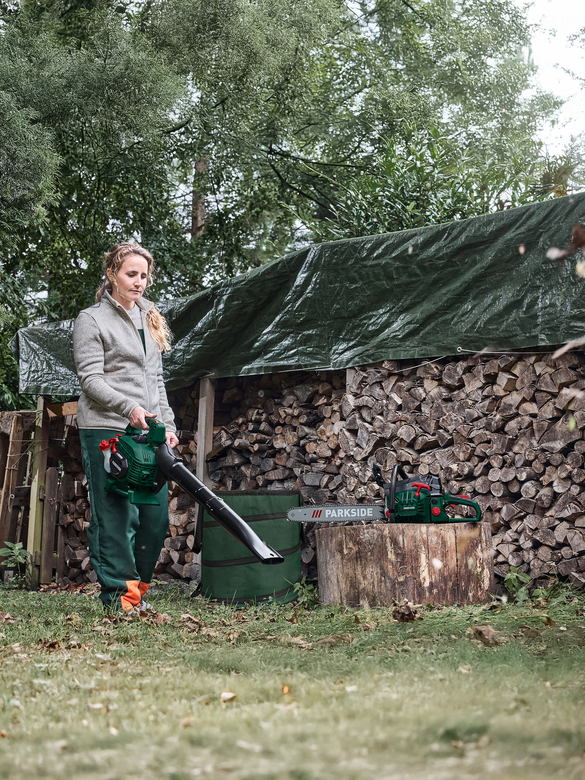 Eine Frau benutzt einen Laubbläser neben einem Holzstapel, mit einer Kettensäge auf einem Baumstumpf.