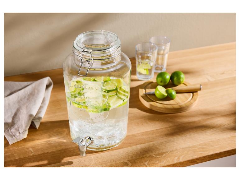 Getränkespender aus Glas mit geschnittenen Limetten, Gläsern und ganzen Limetten auf einem Holztisch.