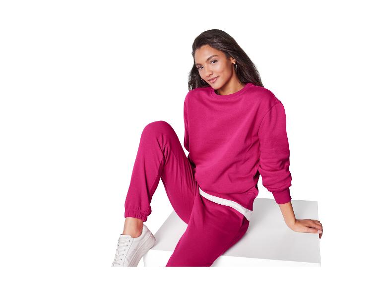 Eine Frau in einem fuchsiafarbenen Sweatshirt und passender Jogginghose sitzt auf einer weißen Oberfläche.