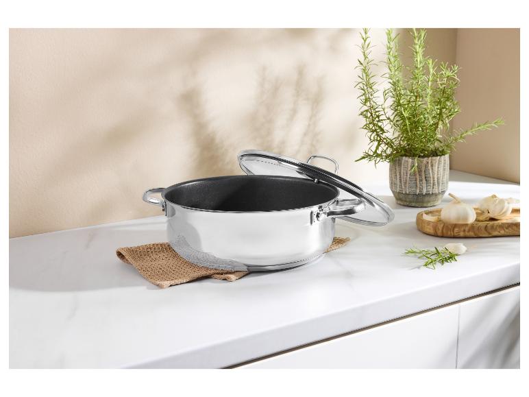 Edelstahl-Kochtopf mit Glasdeckel auf einer weißen Küchenarbeitsplatte mit Kräutern und Knoblauch.