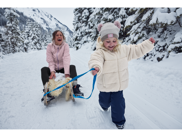 Mutter und Kind in verschneiter Landschaft, das Kind zieht einen Schlitten mit der Mutter.