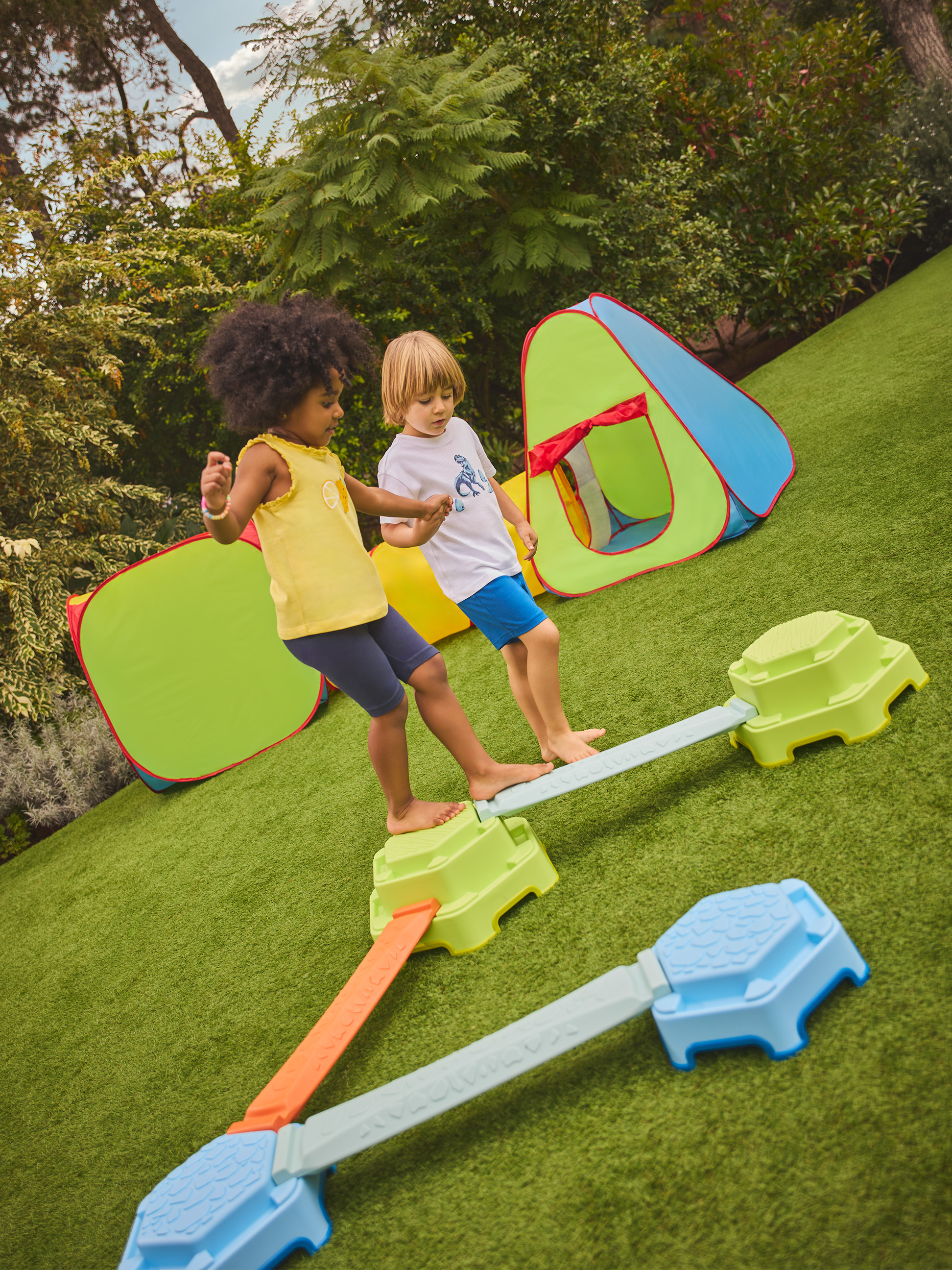 Zwei Kinder spielen auf einem farbigen Balancierpfad mit Spielzelten im Hintergrund.