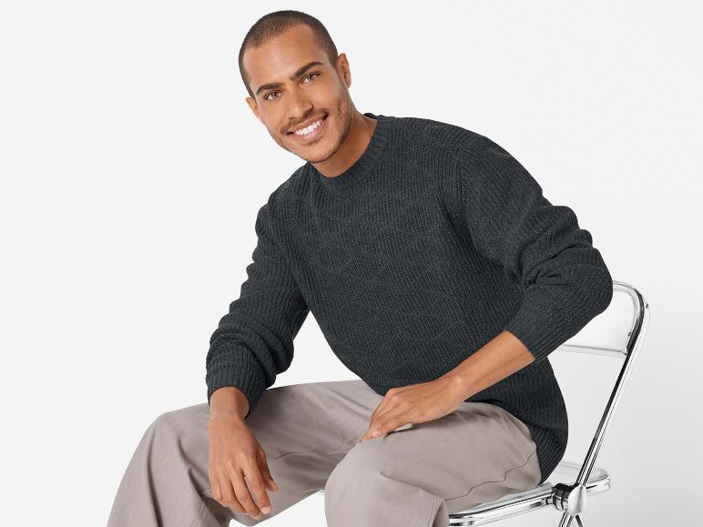 Mann in einem dunkelgrauen Pullover und beigefarbenen Hosen, der auf einem Stuhl sitzt.