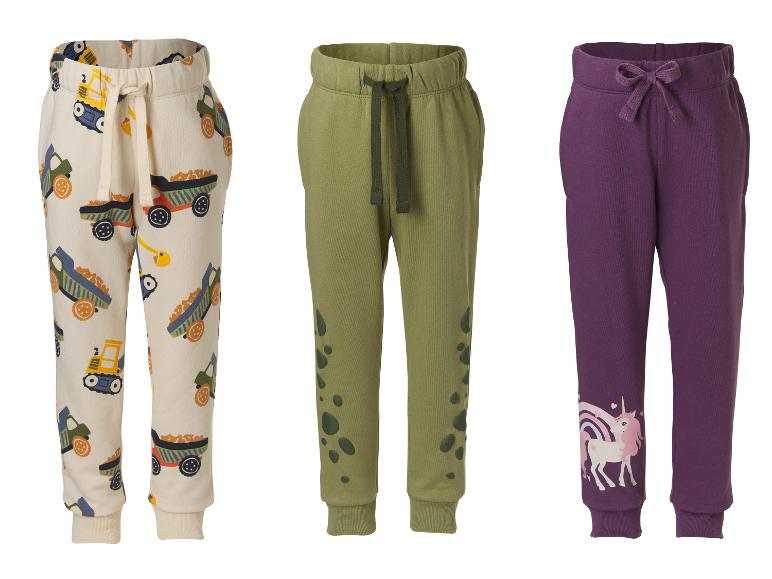 Drei Kinder-Jogginghosen: eine beige mit LKW-Motiven, eine khakigrüne mit Flecken und eine lila mit einem Einhorn.