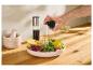 Eine Hand sprüht Öl auf eine Salatschüssel, mit Mörser und Stößel und einer Essigflasche im Hintergrund.