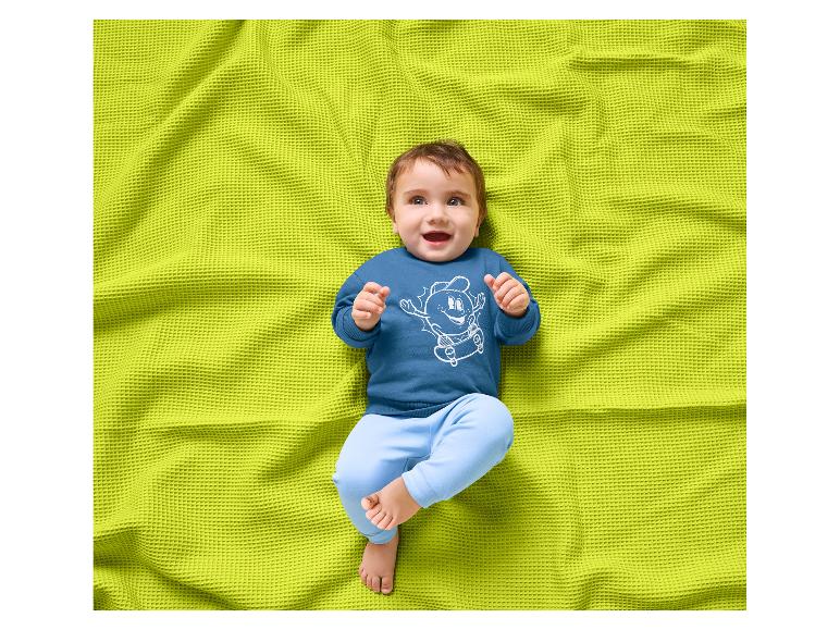 Lächelndes Baby in blauem Sweatshirt mit Skateboard-Motiv und hellblauer Hose auf einer grünen Decke.