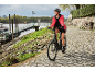 Eine Frau mit Helm und roter Jacke fährt auf einem schwarzen E-Bike auf einem gepflasterten Weg am Fluss.