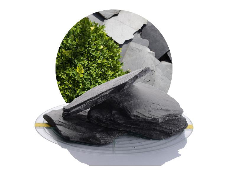 Schiefersteine auf einer Glasplatte, mit einem Busch und weiteren Schieferplatten im Hintergrund.