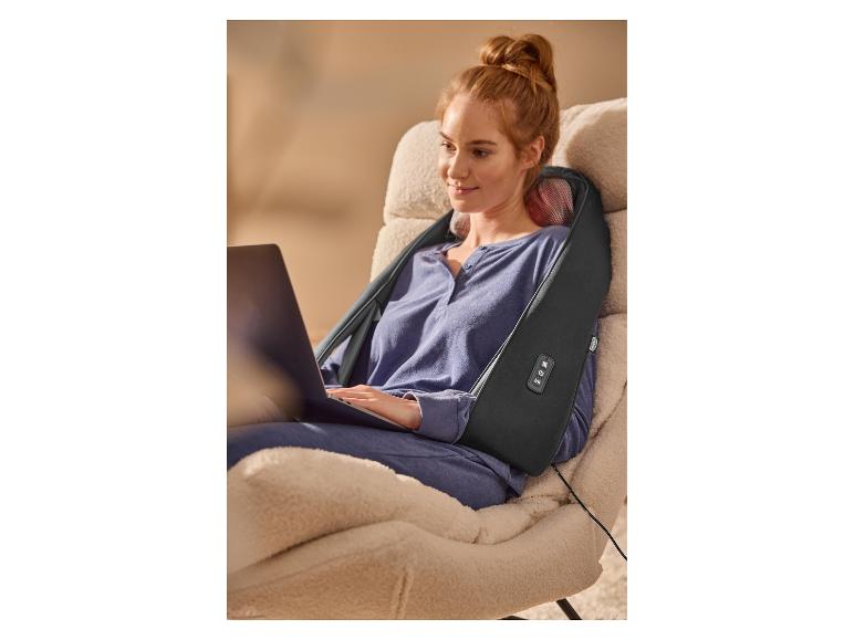 Entspannte Frau in einem Sessel benutzt ein Nacken- und Schultermassagegerät, während sie am Laptop arbeitet.