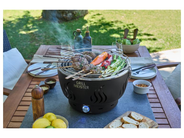 Grill mit Fleisch- und Gemüsespießen auf einem Gartentisch, umgeben von Geschirr und Gewürzen.
