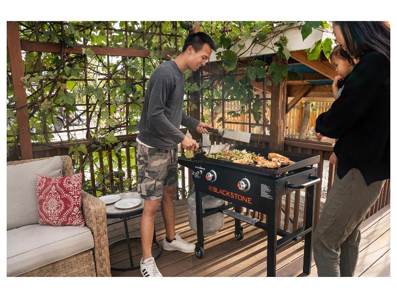 Mann grillt Hähnchen und Gemüse auf einer Grillplatte auf einer Terrasse mit Familie.