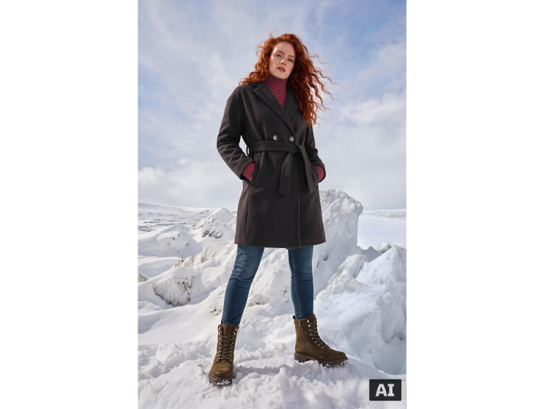 Frau in dunklem Mantel, rotem Rollkragenpullover, Jeans und braunen Stiefeln in Schneelandschaft.