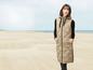 Eine Frau trägt eine lange, beige wattierte Weste mit Kapuze am Strand.