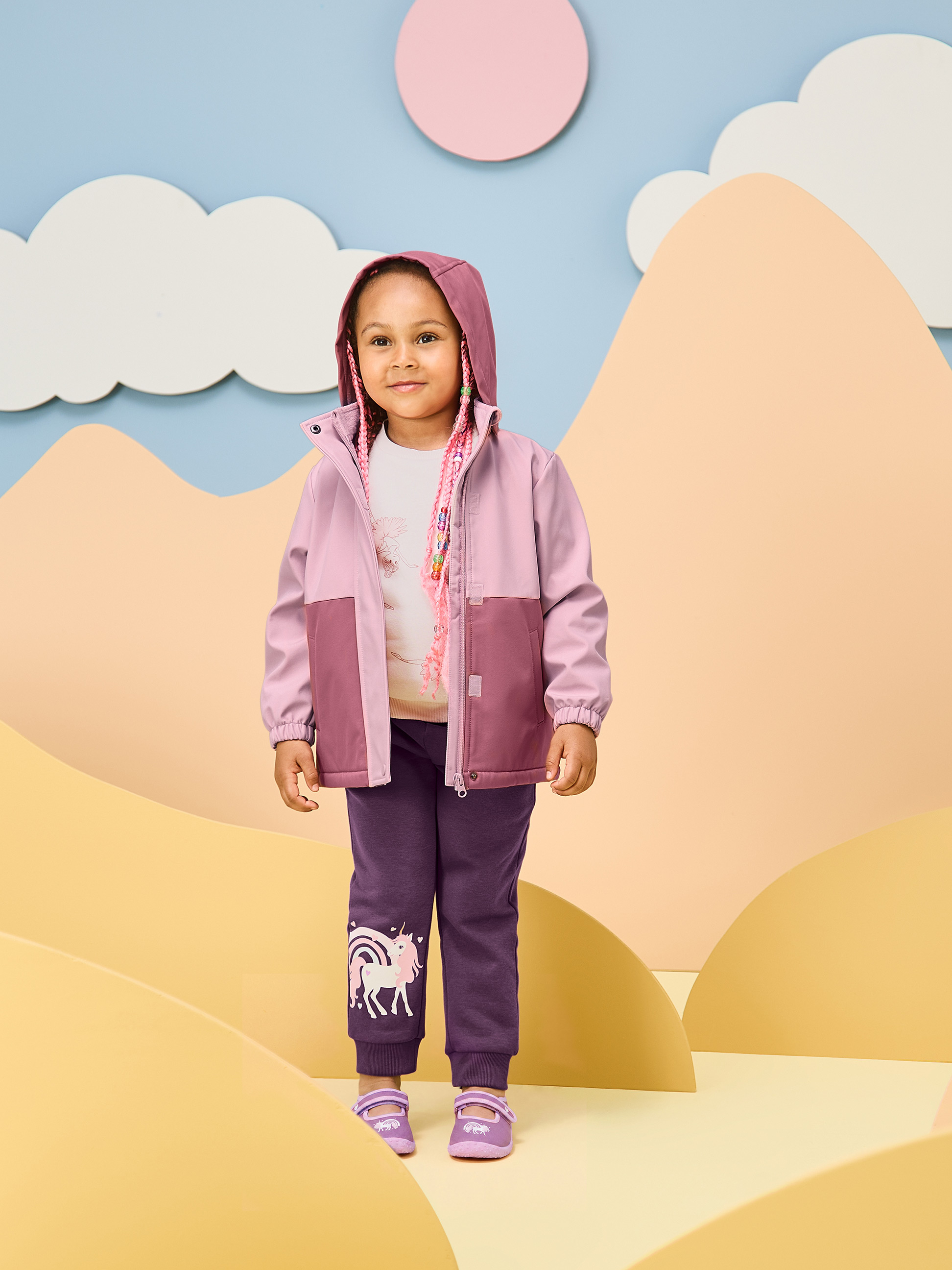 Mädchen in rosa Jacke und lila Einhorn-Hose in spielerischer Umgebung.