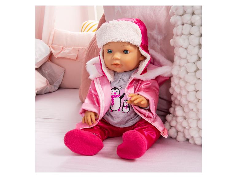 Babypuppe in einem rosa Winteroutfit mit Pinguin-Shirt, auf einem Bett sitzend.