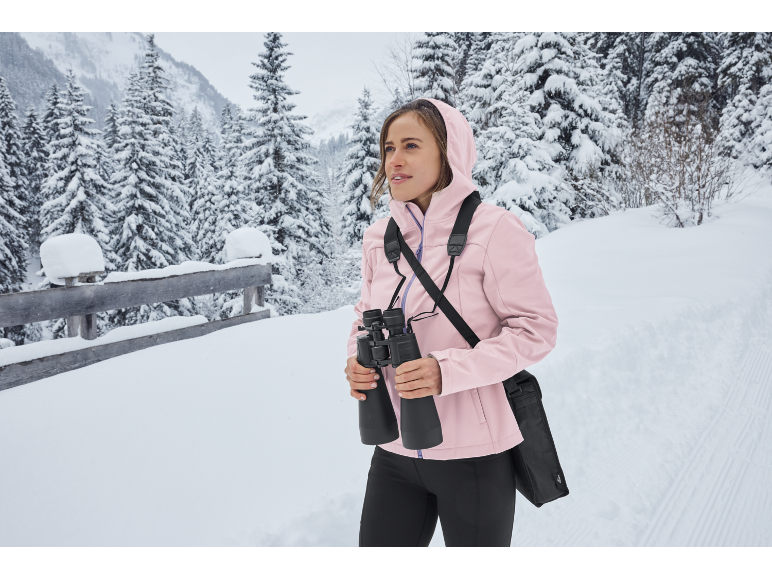 Frau mit Fernglas und rosa Jacke in verschneiter Winterlandschaft.