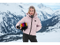 Frau in rosa Skijacke mit einem Helm in der Hand vor verschneiter Bergkulisse.