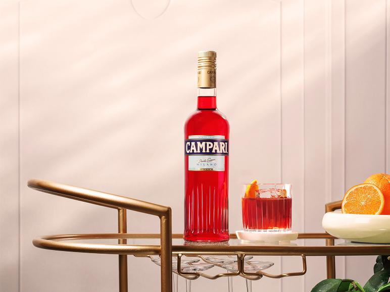 Rote Aperitifflasche und Cocktail mit Orangengarnitur auf einem goldenen Barwagen.