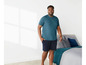 Mann in einem blauen T-Shirt und schwarzen Shorts in einem Schlafzimmer.