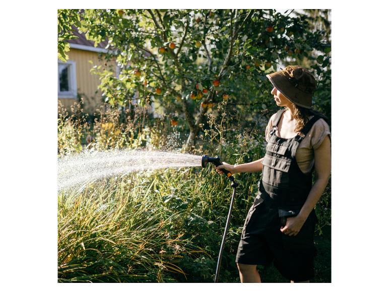 Person in Latzhose und Hut bewässert Garten an einem sonnigen Tag.
