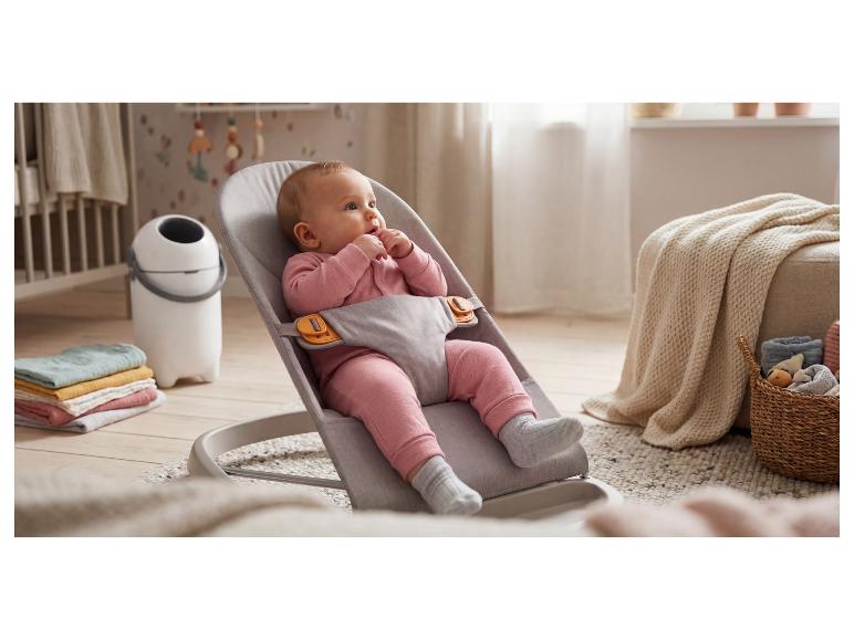 Baby in einer grauen Babywippe, umgeben von Heimtextilien und einem Windeleimer.