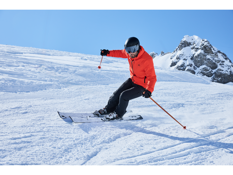 Mann fährt Ski einen verschneiten Hang hinunter, trägt eine rote Skijacke, schwarze Skihose und einen Helm.