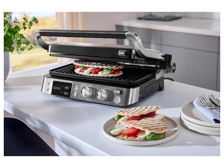 Grill mit zwei Sandwiches, Tomaten, Mozzarella und Basilikum auf einem weißen Tisch.