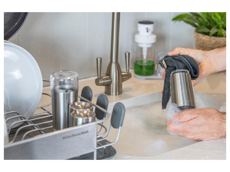 Hände waschen einen Metallbecher in der Küchenspüle mit Seife, nahe einem Abtropfgestell.