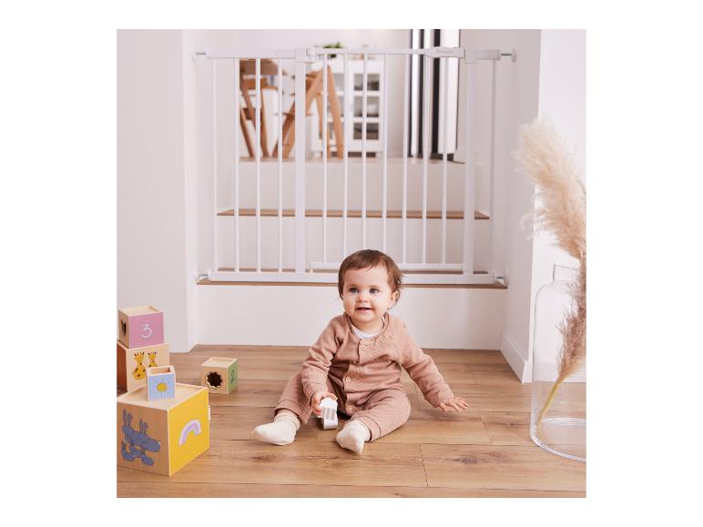 Baby sitzt auf Holzboden mit Spielzeugblöcken und einem weißen Schutzgitter im Hintergrund.