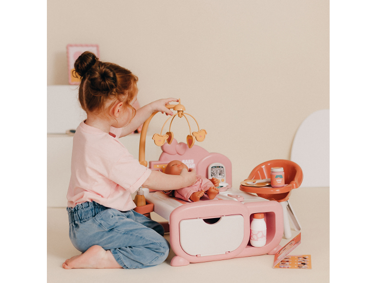 Mädchen spielt mit rosa Puppenpflegeset, inklusive Bettchen und Hochstuhl.