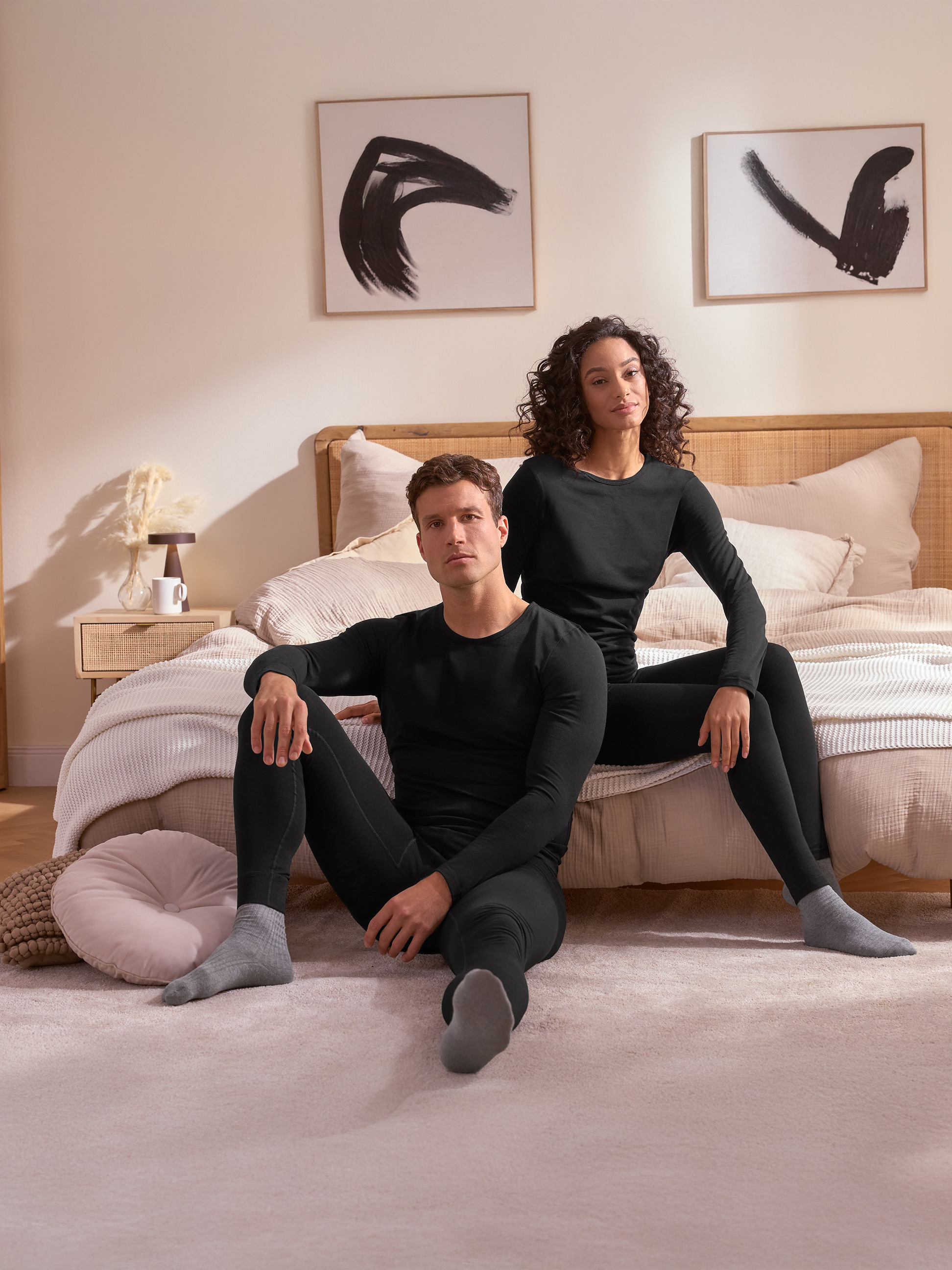 Ein Paar in schwarzer Thermounterwäsche sitzt auf dem Boden vor einem Bett.