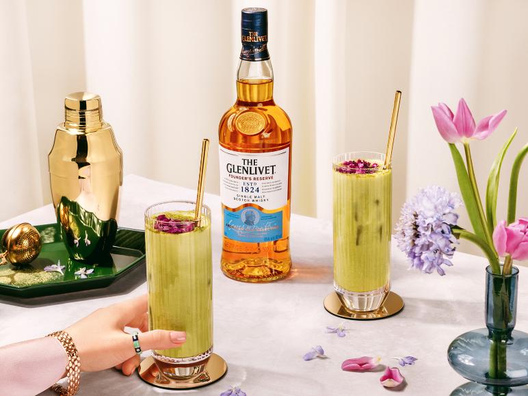 Whiskyflasche und zwei grüne Cocktails mit Rosenblättern, einem goldenen Shaker und Blumen.