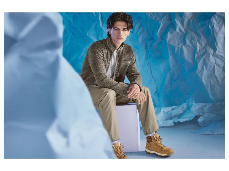 Mann in brauner Jacke und Khakihose, sitzend auf einem weißen Würfel.