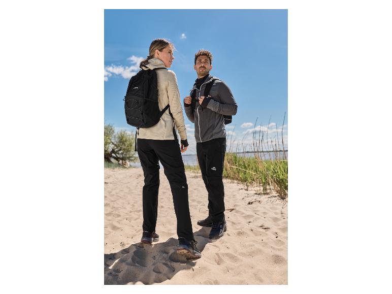 Zwei Wanderer in Outdoor-Kleidung und mit Rucksäcken stehen an einem Sandstrand am Wasser.