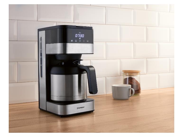 Kaffeemaschine mit Thermoskanne, daneben Kaffeeglas und Tasse auf Holzarbeitsplatte.